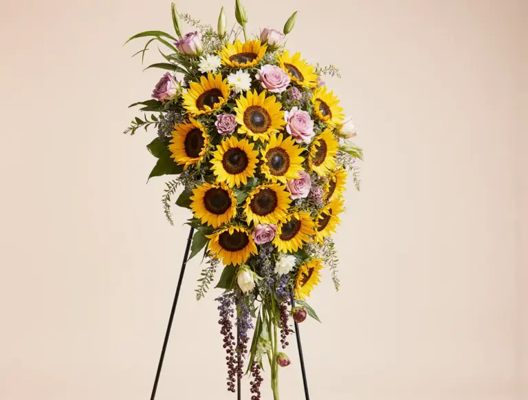 Kukkakauppa Auringonkukkia Liperi palvelut – kaunis hautajaisiin tarkoitettu kukkalaite auringonkukista, ruusuista ja vihreistä oksista jalustalla.